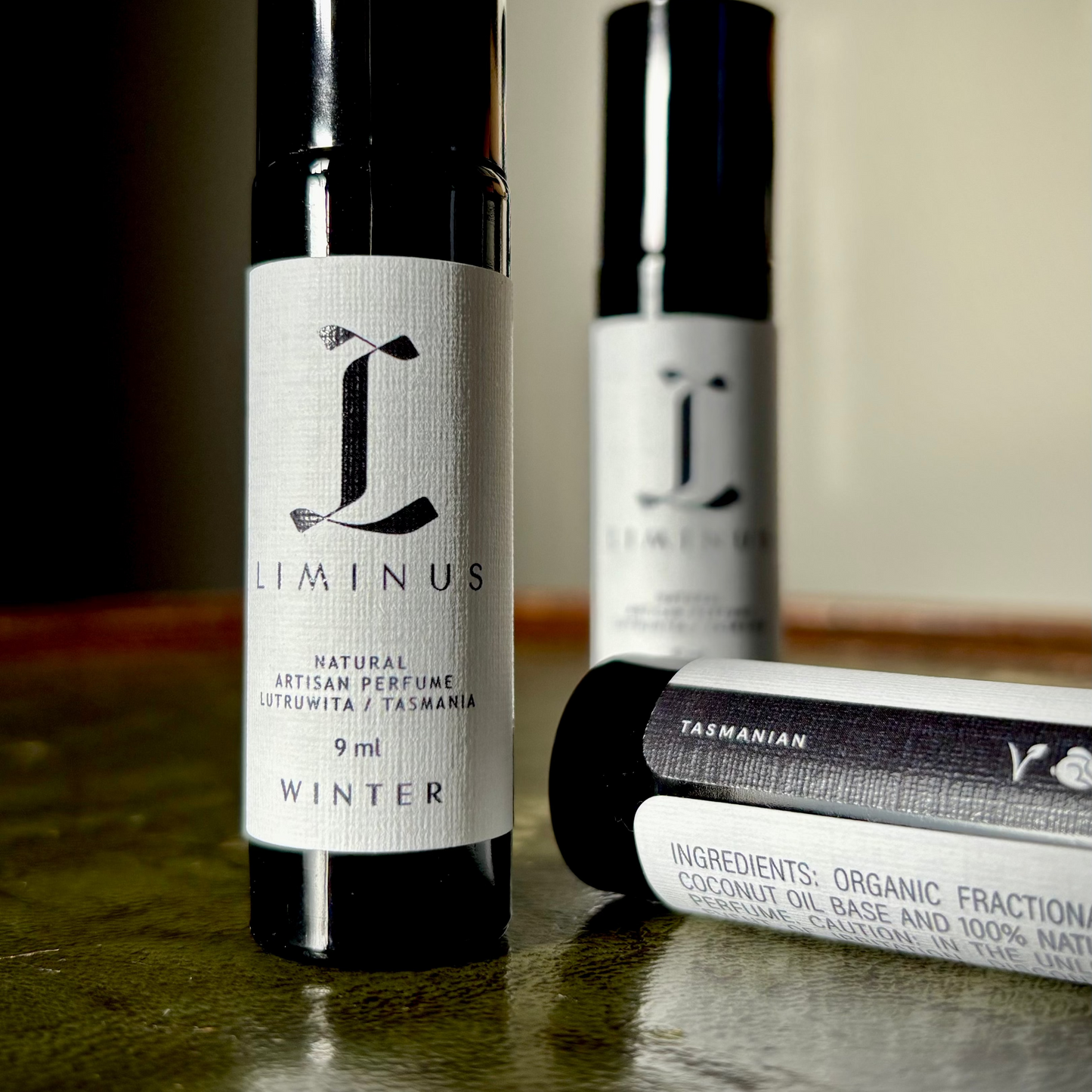 Three black cylindrical perfume bottles with white labels from the brand Liminus, arranged on a dark green leather surface against a warm beige background. One bottle stands upright in the foreground, another stands blurred in the back, and a third lies on its side showing product details.